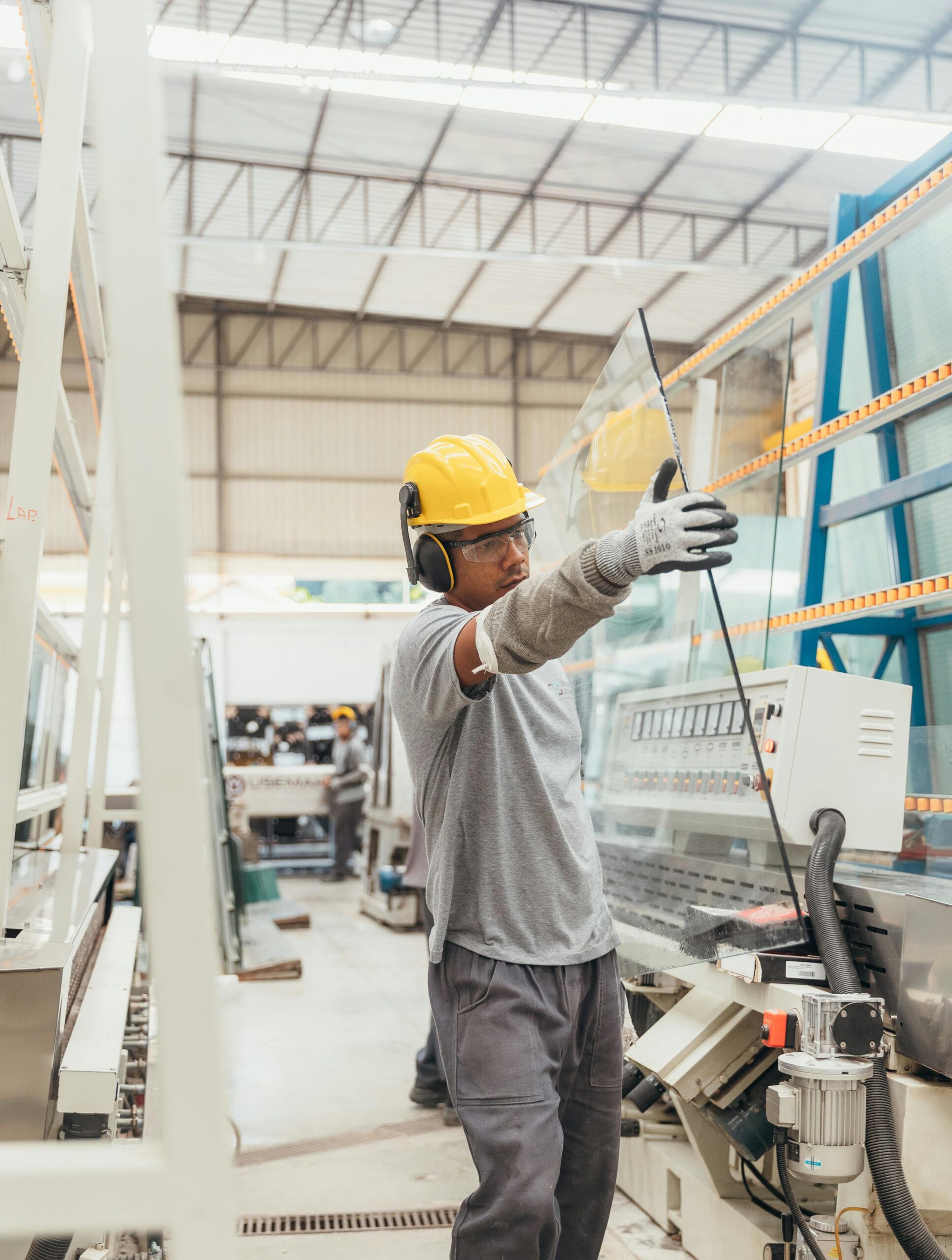 Man carrying a sheet of glass wearing a hardhat, safety glasses, earmuffs, and gloves in a warehouse.